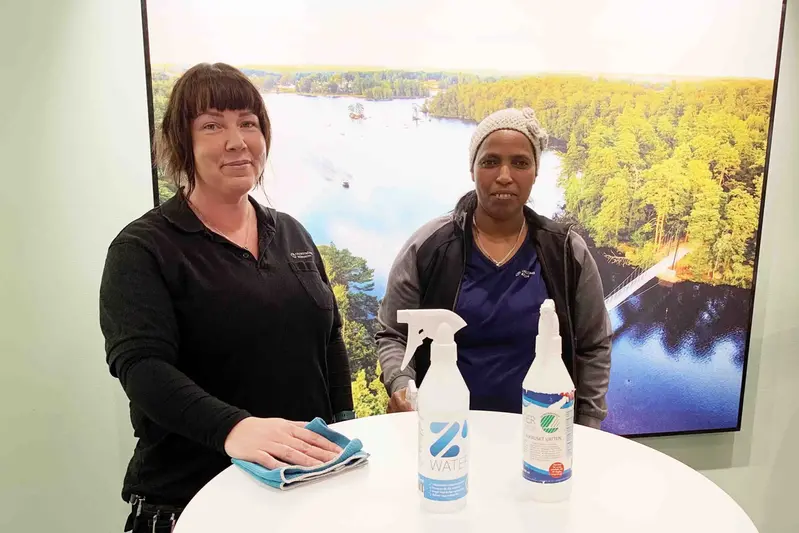 Två kvinnor anställda som lokalvårdare i Olofströms kommun står framför ett bord med två sprayflaskor med rengöringsmedel. 
