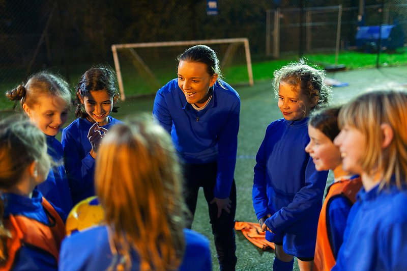 En vuxen står framåtböjd med sju flickor i en ring på en fotbollsplan